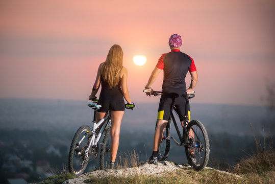 Rear view cyclist couple with mountain bikes standing on a rock, enjoying the evening scenery. Bright sun in between. Blurred background