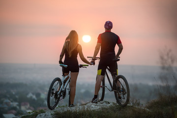 Naklejka premium Rear view cyclist couple with mountain bikes standing on a rock holding hands enjoying the sunset. Bright sun in between. Blurred background