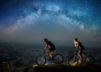 Obraz premium Sporty friends riding a bicycles on the mountain hill at night under starry sky and Milky way. Below is a city in the distance
