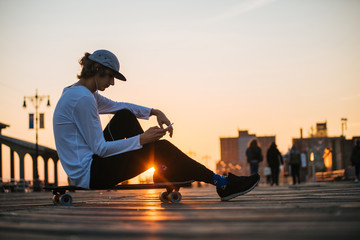 Young hipster man silhouette sitting on the longboard using his phone and headphones