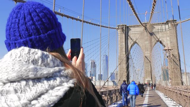Young female tourist taking photos with her smart phone on Brooklyn Bridge NYC