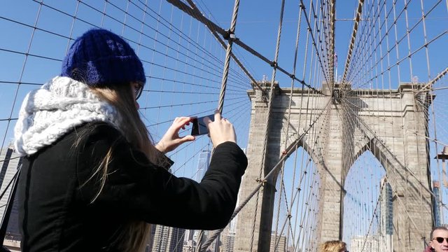 Young female tourist taking photos with her smart phone on Brooklyn Bridge NYC
