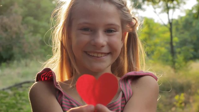 Happy smiling child kid girl holding red paper heart in her hands against warm sunny nature background. Mother's Day, Valentine's day, love nature or childhood happiness concept