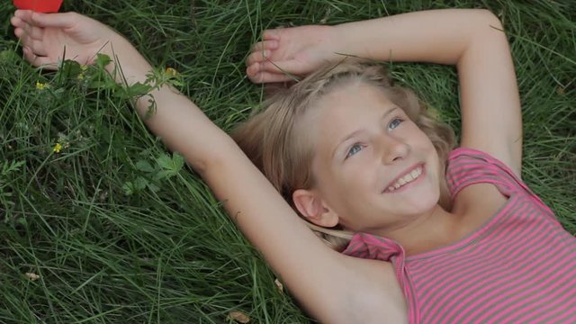 Happy smiling young girl lying on grass with arms outstretched having fun holding red paper heart in her hand. Love nature or happiness childhood concept