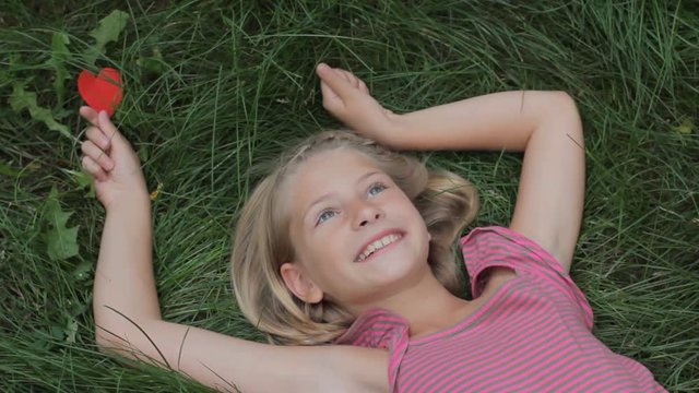 Happy smiling kid girl lying on grass with arms outstretched holding red paper heart in her hand. Love nature or happiness childhood concept