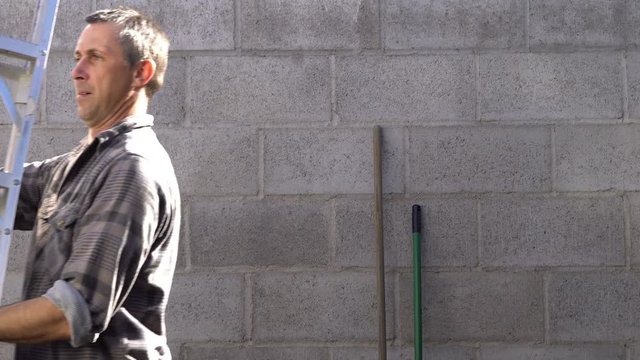 Outdoor shot of a man walking into the shot and picking up a stepladder and then carrying it off to do some house work or home repairs.