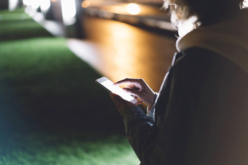 Girl pointing finger on screen smartphone on background illumination bokeh color light in night city, hipster using in hands and texting mobile phone, mockup glitter street, content lifestyle