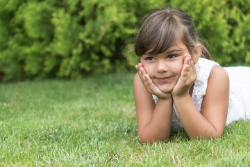 Cool brunette little girl lying on the grass supporting her chin with both hands is looking ahead. Free place for your text is in the left side of the photo.