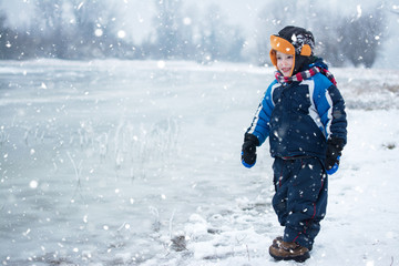 Cute little boy standing near frozen lake during cold snowy day.