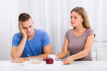 Young wife is giving healthy breakfast to her husband but he doesn't like it.
