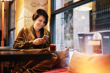 Beautiful caucasian female with dark hair is typing messages on her smartphone while sitting beside the window of a cozy and modern coffee shop and drinking rea during the free time.