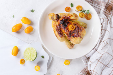 Whole roasted mini chicken cooking with caramelized kumquats served on white plate with kitchen towel, cutting boards and fresh citrus fruits over white concrete texture background. Top view