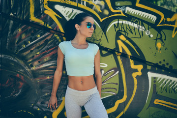 Young attractive woman with sunglasses standing against graffiti wall in park and posing after fitness activity.