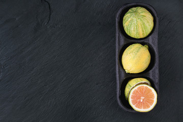 Obraz na płótnie Canvas whole and sliced citrus fruit pink tiger lemonin paper market box over black stone slate textured background. Top view with space. Modern healthy eating