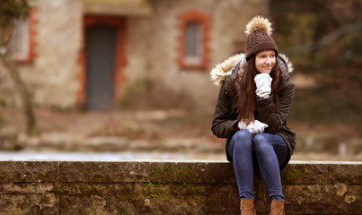 Glückliche junge modisch gekleidete Frau im Winter sitzt auf einer Mauer