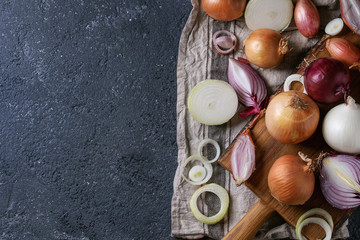 Variety of whole and sliced red, white, yellow and shallot onions on wooden chopping cutting board on textile napkin over dark stone texture background. Top view, space for text
