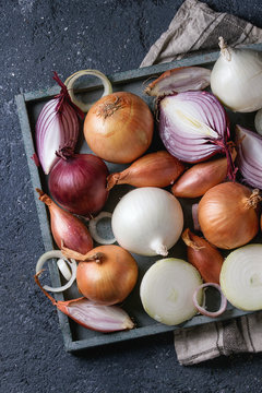Variety of whole and sliced red, white, yellow and shallot onions on wooden square tray on kitchen towel over dark stone texture background. Top view, space for text