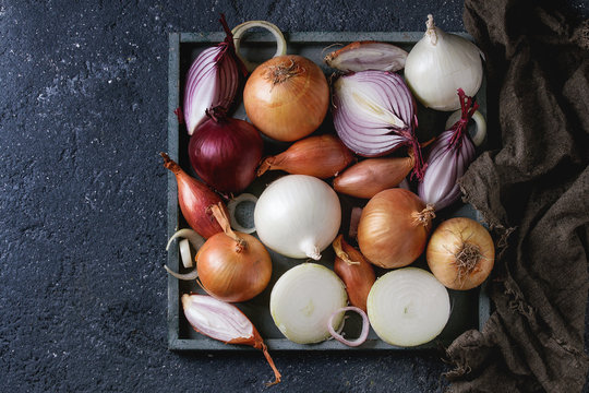 Variety of whole and sliced red, white, yellow and shallot onions on wooden square tray on sackcloth rag over dark stone texture background. Top view, space for text