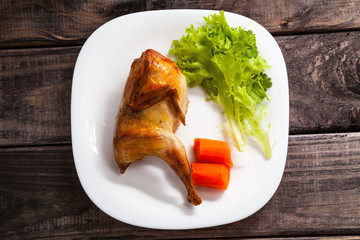 Roasted quail on a white ceramic plate. Serving with carrot and salad lettuce on old dark grey rustic wooden table. Top view. Dinner menu.
