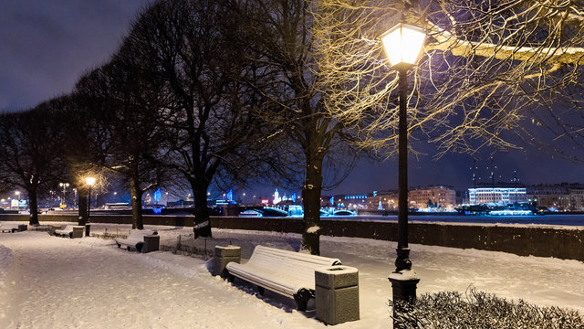 Benches under snow along alley with trees and street lights on embankment. Winter cityscape at twilight. Saint Petersburg, Russia. Selective focus.