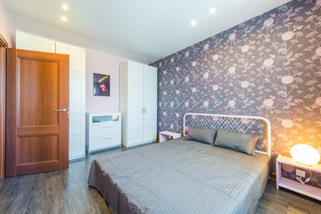 Interior bedroom with a large double bed with bedside tables,wardrobes, chest of drawers on a background of modern wallpaper