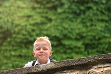 Blonder Junge im Anzug schaut lächelnd über eine Mauer