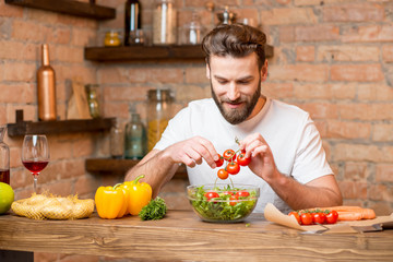 Handsome bearded man in white t-shirt making salad with tomatoes and pepper in the kitchen. Healthy and vegan food concept