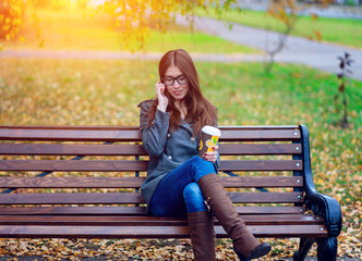 Girl in jacket talking on the phone, sitting  a bench, holding  coffee or tea,  young outdoors, spring  fall, life style, the concept of the city, lifestyle, listening to the conversation.