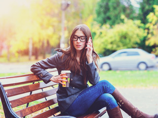 Girl in jacket talking on the phone, sitting  a bench, holding  coffee or tea,  young outdoors, spring  fall, life style, the concept of the city, lifestyle, listening to the conversation.