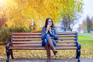 Beautiful young woman sitting on a bench drinking coffee or hot tea in the spring  autumn coat enjoying in park outdoors, glasses, urban life, the concept of breakfast in nature - Powered by Adobe