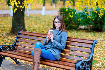 Beautiful young woman sitting on a bench drinking coffee or hot tea in the spring  autumn coat enjoying in park outdoors, glasses, urban life, the concept of breakfast in nature
