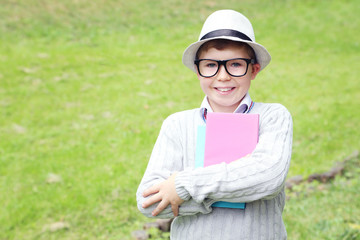 Fototapeta premium Portrait of young boy with eyeglasses, books and hat in the park