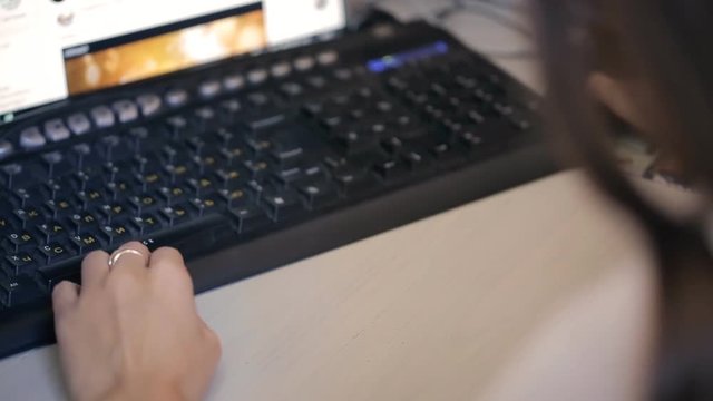 Black keybord in front of monitor with one woman hand. There is view from persons back on standard keypad with yellow Russian letters, white English alphabet, grey special buttons on top. It is on
