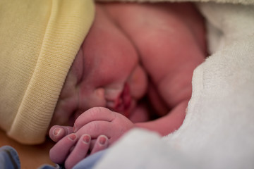 Newborn baby hands clasping together with fresh born face in soft focus pouting lips asleep skin to skin with maternal mother swaddled up tight