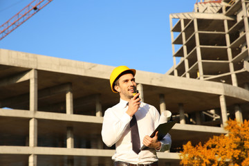 Handsome engineer with portable radio transmitter and clipboard standing against unfinished building