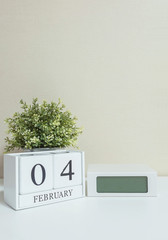 White wooden calendar with black 4 february word with clock and plant on white wood desk and cream wallpaper textured background in selective focus at the calendar