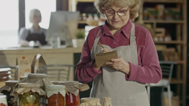 Tilt up of elderly female owner in glasses standing in farmer market and counting canned produce 