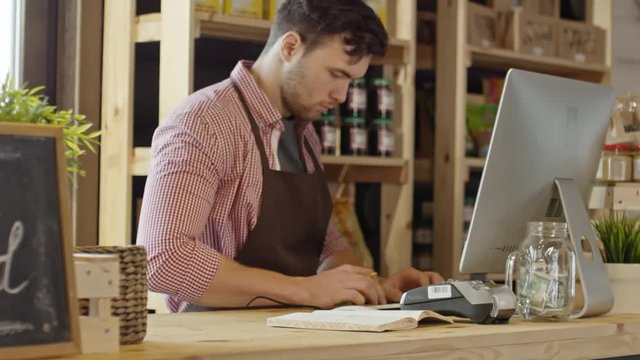 Panning shot of young hipster man in apron standing behind counter in organic food shop typing something on computer and writing notes in notepad 