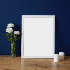 Empty modern style frame, bouquet of white flowers ranunculus and two white candles on a deep blue wall background and wooden table. Poster mockup in hipster romantic style interior