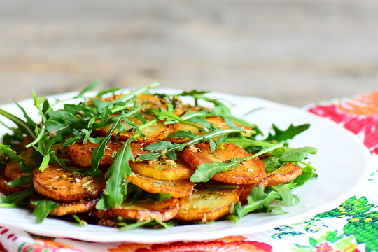 Fried sliced potato with fresh arugula and spices on a white plate on a wooden table. Combine fresh arugula mixture and hot potatoes. Tasty and healthy recipe. Closeup