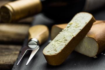 Fototapeta premium Chunk and thick cut off slice of smoked goat cheese with fork on black stone board, bottle of wine in the background, gourmet meal, close up