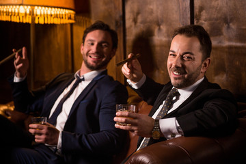 Rich businessmen resting and relaxing in men's club. Happy men drinking whiskey and smoking cigars. People looking at camera.