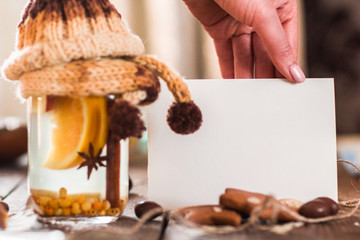 Blank card with hot punch closeup. Female hand holding blank card near glass jar with warm winter drink of orange, cinnamon and anise, decorated with knitted cover, free space