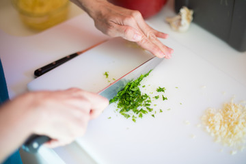 woman hands chopping green fresh parsley after garlic with big kitchen knife on white cutting board
