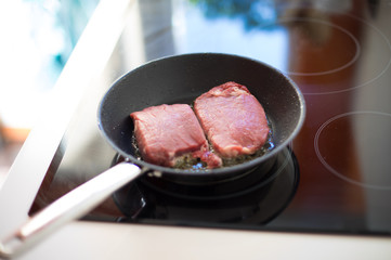 silver metal dripping or frying pan or griddle cooking fresh beef sirloin steaks on black cooker in white kitchen
