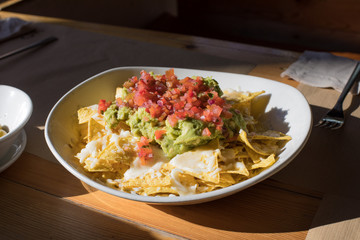 white big bowl with nachos and guacamole, chopped tomato and cheese, on wooden table at restaurant
