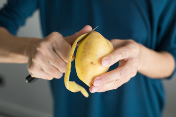 detail of hands of woman with blue sweater peeling fresh yellow potato with kitchen knife,...