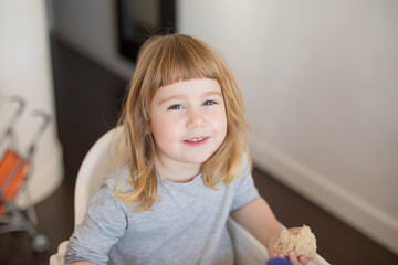 Three years old blonde child with grey shirt eating bread and looking, sitting in white chair indoor
