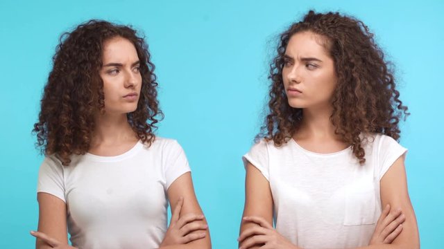 Two grumpy resentful young Caucasian female twins in white t-shirts standing close to each other with arms folded on blue background