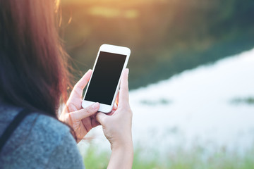 Mockup image of a woman using smart phone with blank black screen at outdoor and lake nature background